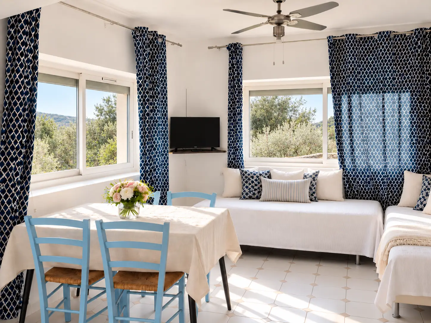 Pièce de vie du gîte Les Figuiers avec table et chaises bleues, banquettes claires et fenêtres sur la végétation